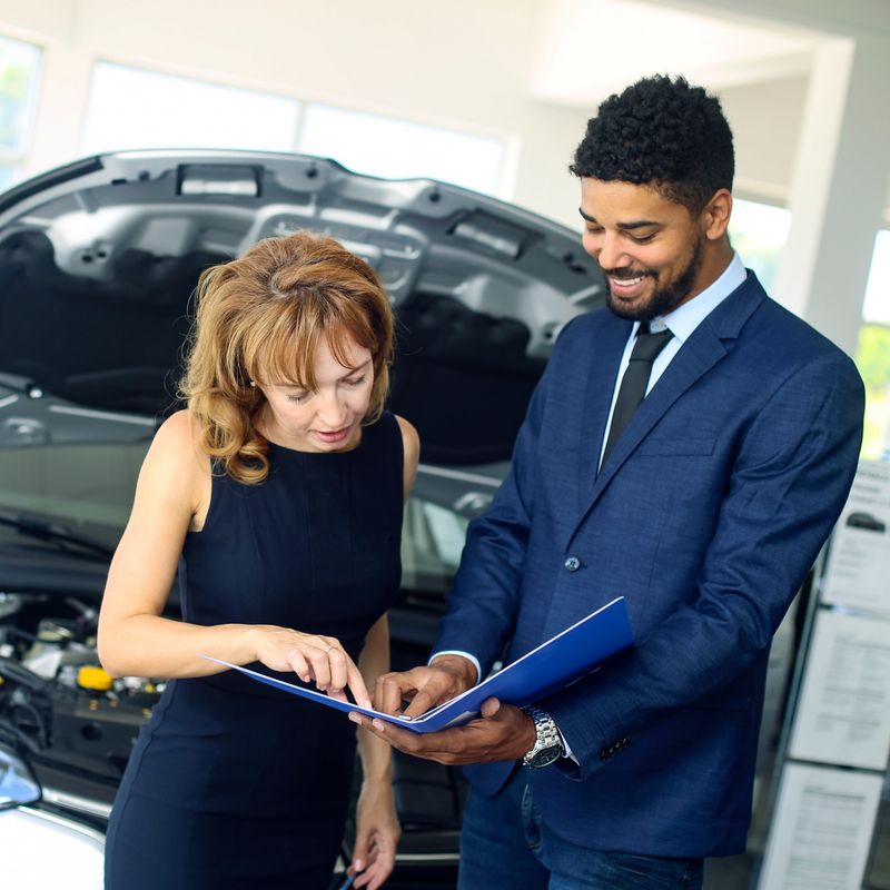 Salesman and a customer in a car dealership. Mixed race male and a blonde female.