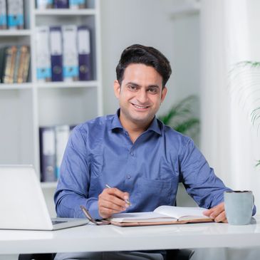 Man in blue shirt smiling while working at a desk with a laptop and notebook.