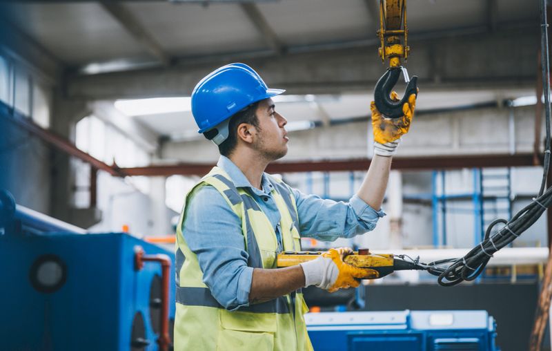 Young heavy industry worker man holding crane hook button working with safety workwear and moving a massive metal construction object in warehouse of factory. XXXL size