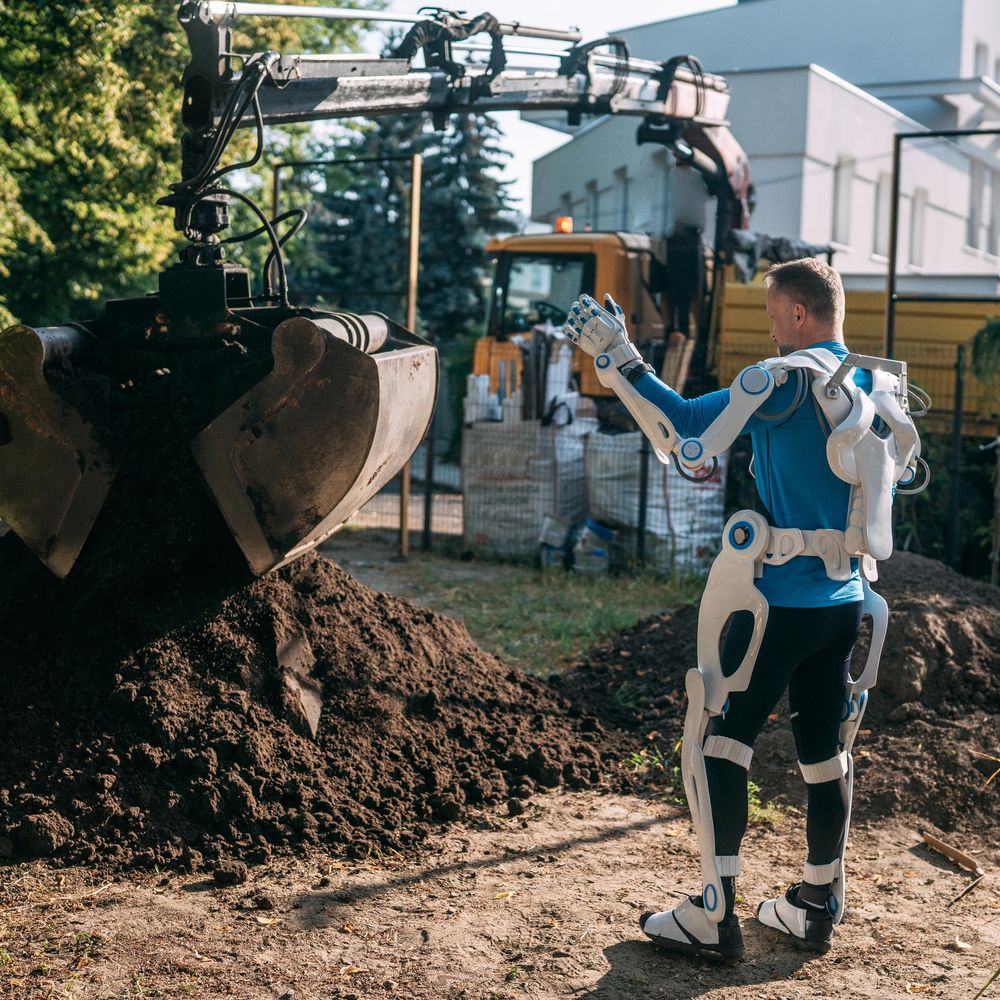 Man wearing a robotic exoskeleton controlling heavy excavation machinery outdoors.