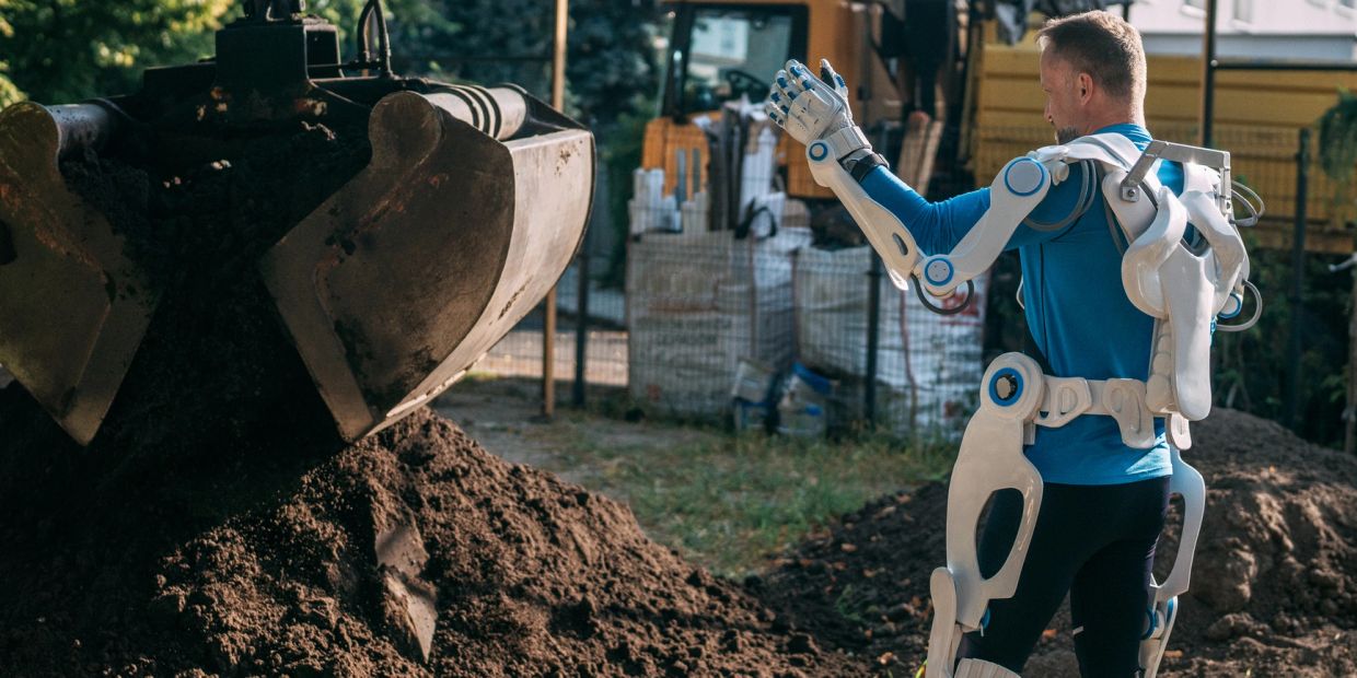 Man wearing robotic exoskeleton working with excavator outdoors.