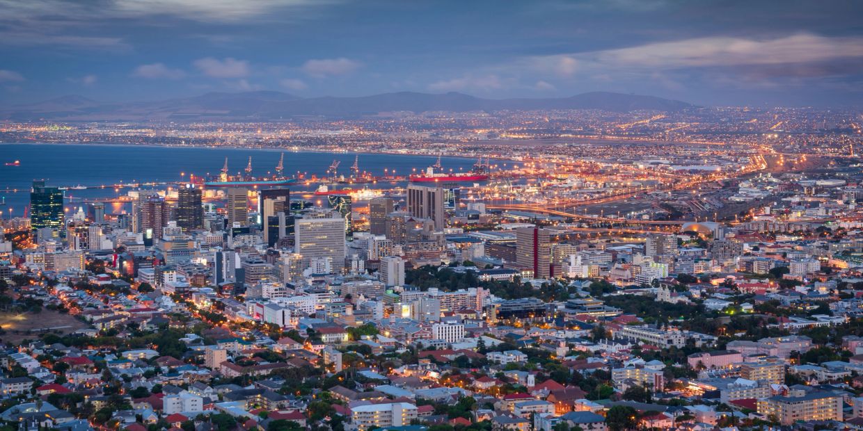 Cityscape of a coastal city at dusk with illuminated buildings and harbor.