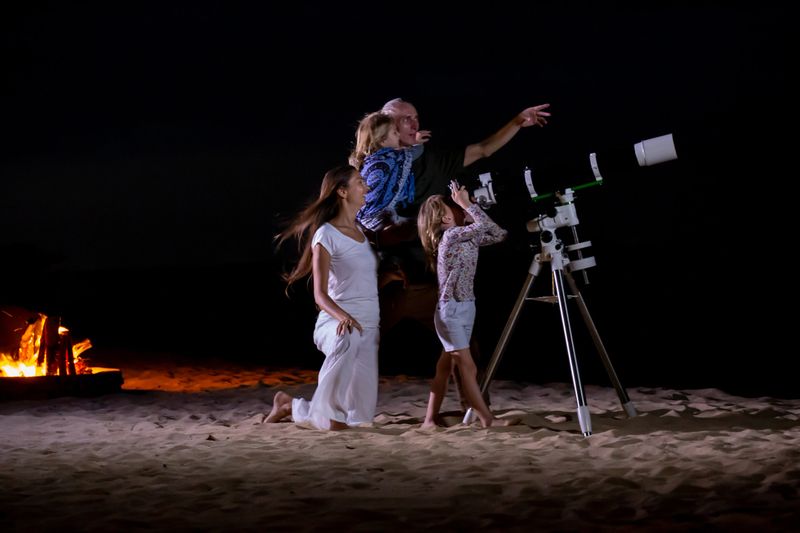 Happy family looking at moon and stars through a telescope at beautiful night sky. Parents and children star gazing on the beach