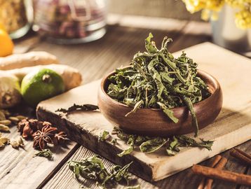 Dried herbs in a wooden bowl surrounded by spices and fresh ingredients on a rustic wooden surface.