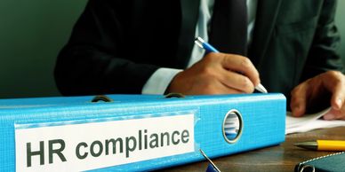 A blue binder labeled HR compliance on a desk with a person writing in the background.