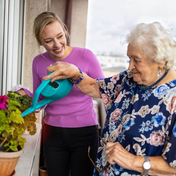 Young woman helps elderly woman water plants on balcony.