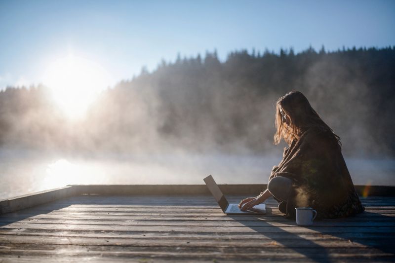 Side view of beautiful woman relaxing at sunrise with coffee and using laptop.