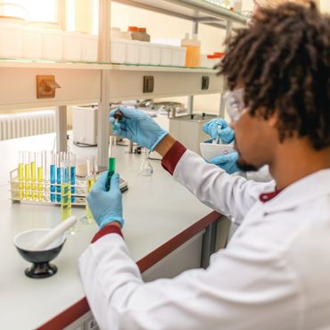 Scientist in a lab adding liquid to a test tube, wearing gloves and safety goggles.