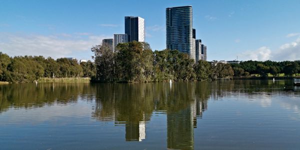 City buildings reflected in a calm lake surrounded by trees.