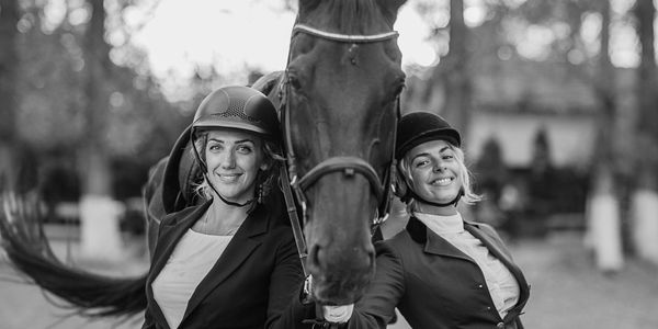 Two women in equestrian helmets posing with a horse, smiling outdoors.