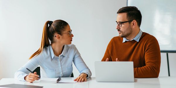 Two colleagues discussing work with a laptop and notebook in a modern office.