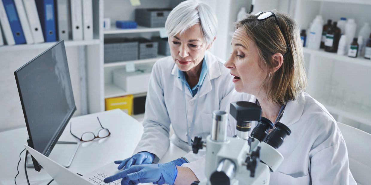 Two female scientists discussing data in a lab with a microscope and computer.