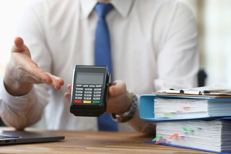 Close-up of business male holding cashier electrical machine for fast payment. Nfc technology counter. Man in suit with folder on table. Transaction and pay bill on terminal