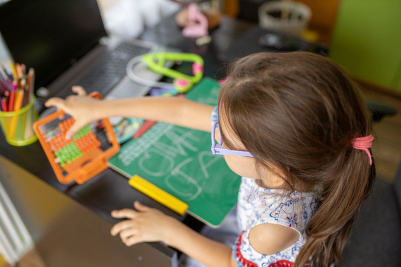 Beautiful young girl with eyeglasses is sitting at her desk and drawing on a board. Preschool child.