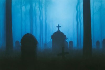 Foggy graveyard with silhouetted tombstones and crosses amid tall trees at dusk.