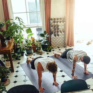 Two people doing push-ups on yoga mats in a bright, plant-filled room.
