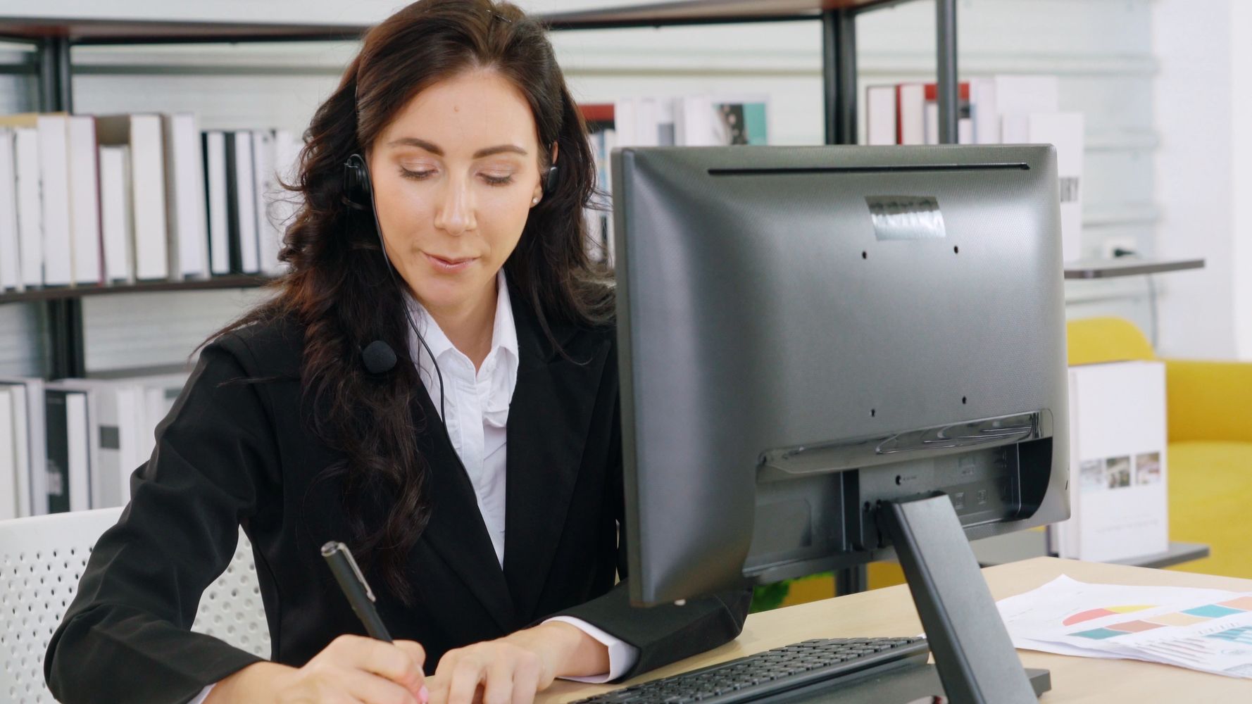 Woman in headset working at computer and writing notes in office.