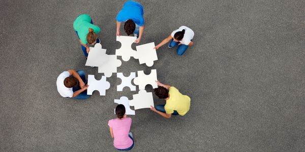 Six people collaborating to assemble oversized puzzle pieces outdoors.