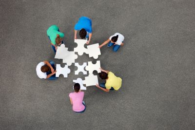 Six people collaboratively assembling large white puzzle pieces on gray ground.