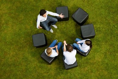 Four people sitting on black cube seats in a circle on grass, engaged in conversation.