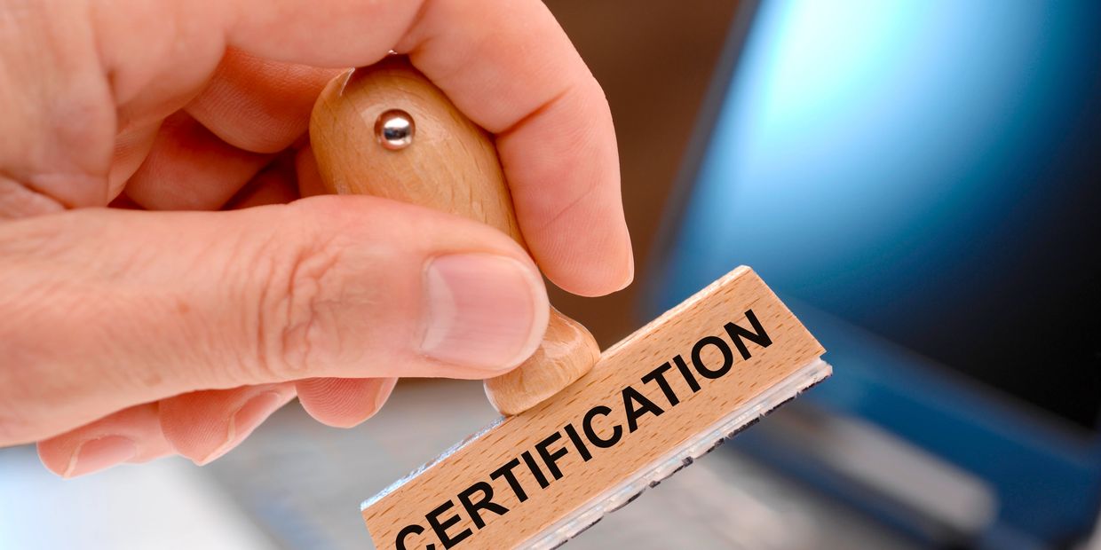 Hand holding a stamp labeled 'CERTIFICATION' with a laptop in the background.