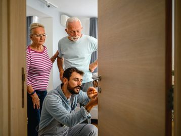 Young technician fixing a door lock while elderly couple watches.