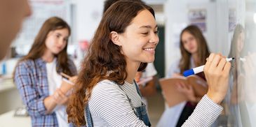 Young woman writing on whiteboard as classmates observe and take notes.