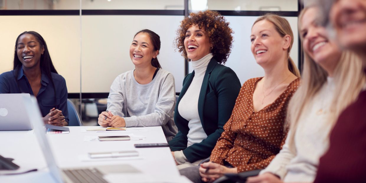 A group of diverse women smiling and engaged in a meeting around a conference table.
