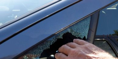 Hand reaching through a broken car window with shattered glass.