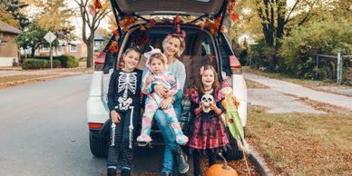 A family dressed in Halloween costumes posing by a car with autumn decorations.