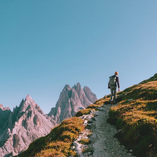 A hiker walks up a mountain trail with jagged peaks in the background.