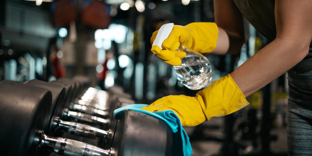 Person cleaning gym dumbbells with spray and cloth wearing yellow gloves.