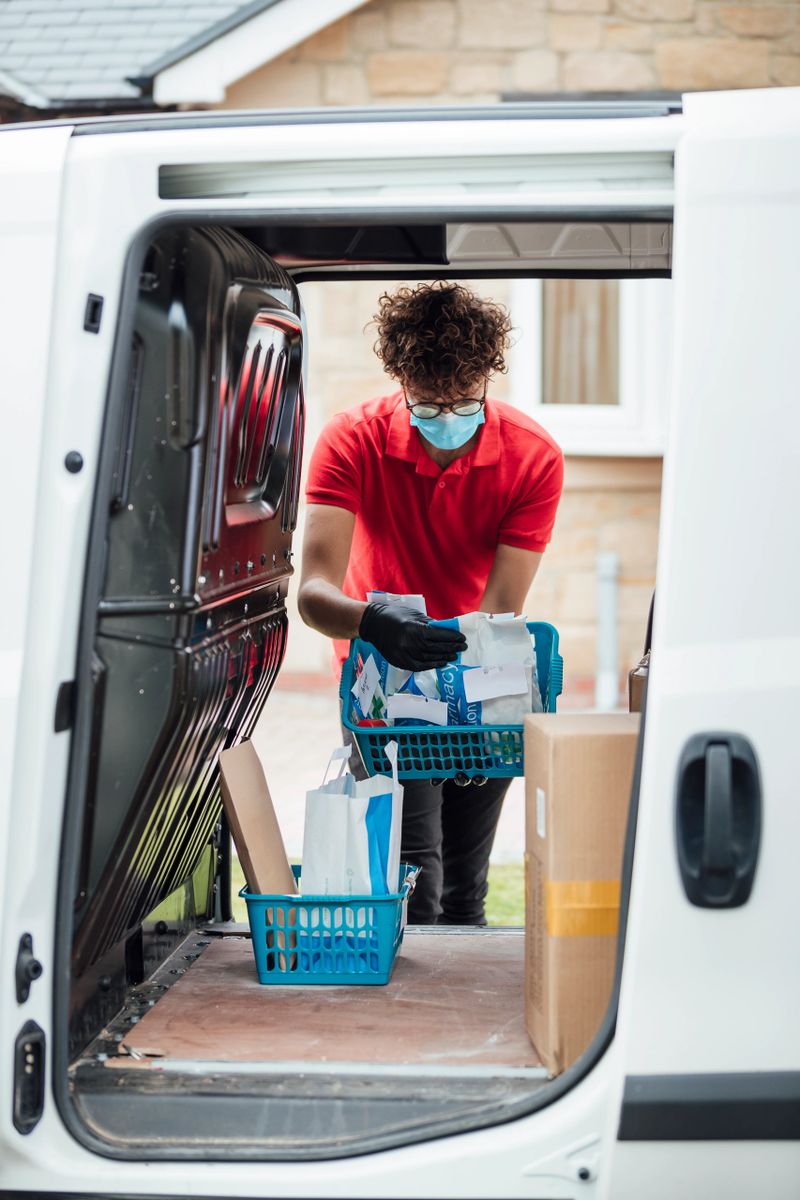 A shot through a van of a keyworker, wearing a protective face mask, holding a basket of prescription medicine. He is looking at where to deliver next.