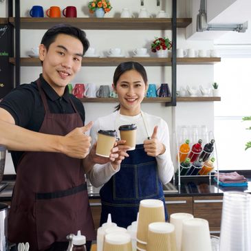 Two baristas in aprons smiling and holding takeaway coffee cups.
