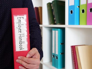 Person holding a red Employee Handbook binder in an office setting.