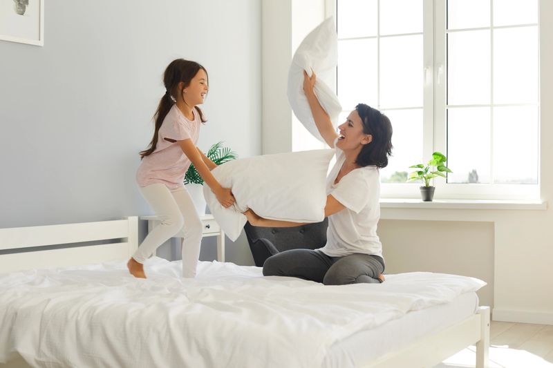 Happy mother and cheerful little daughter having pillow fight on wide bed. Young mommy and cute child fighting with soft white pillows and laughing enjoying weekend together at home