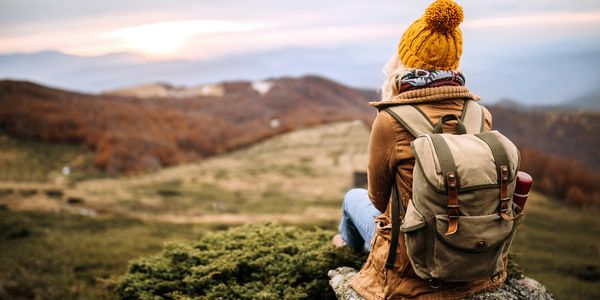 Person in a yellow beanie sitting on a rock, overlooking a scenic autumn landscape.