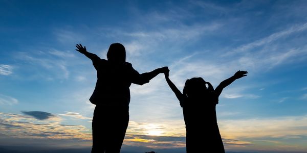 Silhouettes of a woman and child holding hands with arms raised against a sunset sky.