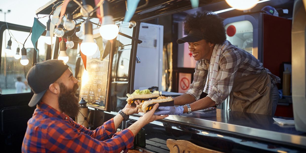 A smiling vendor hands hot dogs to a happy customer at a food truck during sunset.