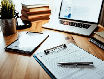 Workspace with tax form, pen, tablet, laptop, books, and plant on wooden desk.