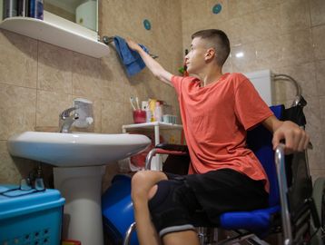 A boy in a wheelchair cleaning a bathroom mirror.