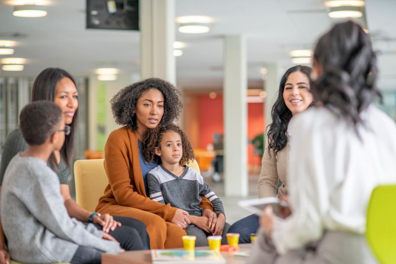 Women and kids sit around a table in a colourful facility and discuss.