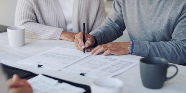 A person signing a document at a table with another person nearby.