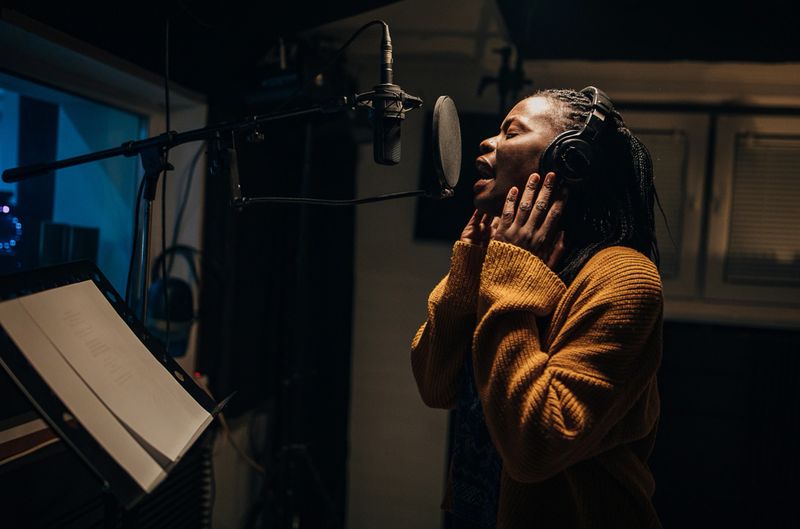 One woman, beautiful black female vocalist wearing headphones and singing into microphone in recording studio.