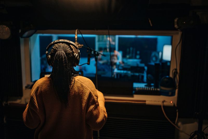 Two people, beautiful black female vocalist wearing headphones and singing into microphone in recording studio, man sound engineer working at mixing panel.