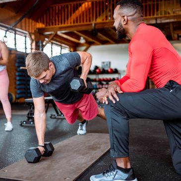 A personal trainer coaching a man lifting dumbbells while a woman exercises with a kettlebell.