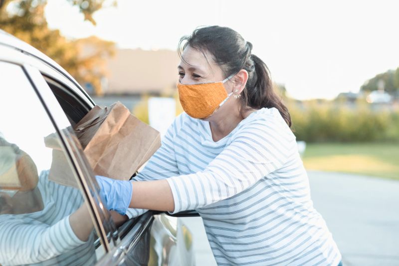 At a food drive during COVID-19, a mature adult woman cheerfully hands a paper bag full of food through the window of a car.  She is wearing a protective mask and gloves to flatten the curve.