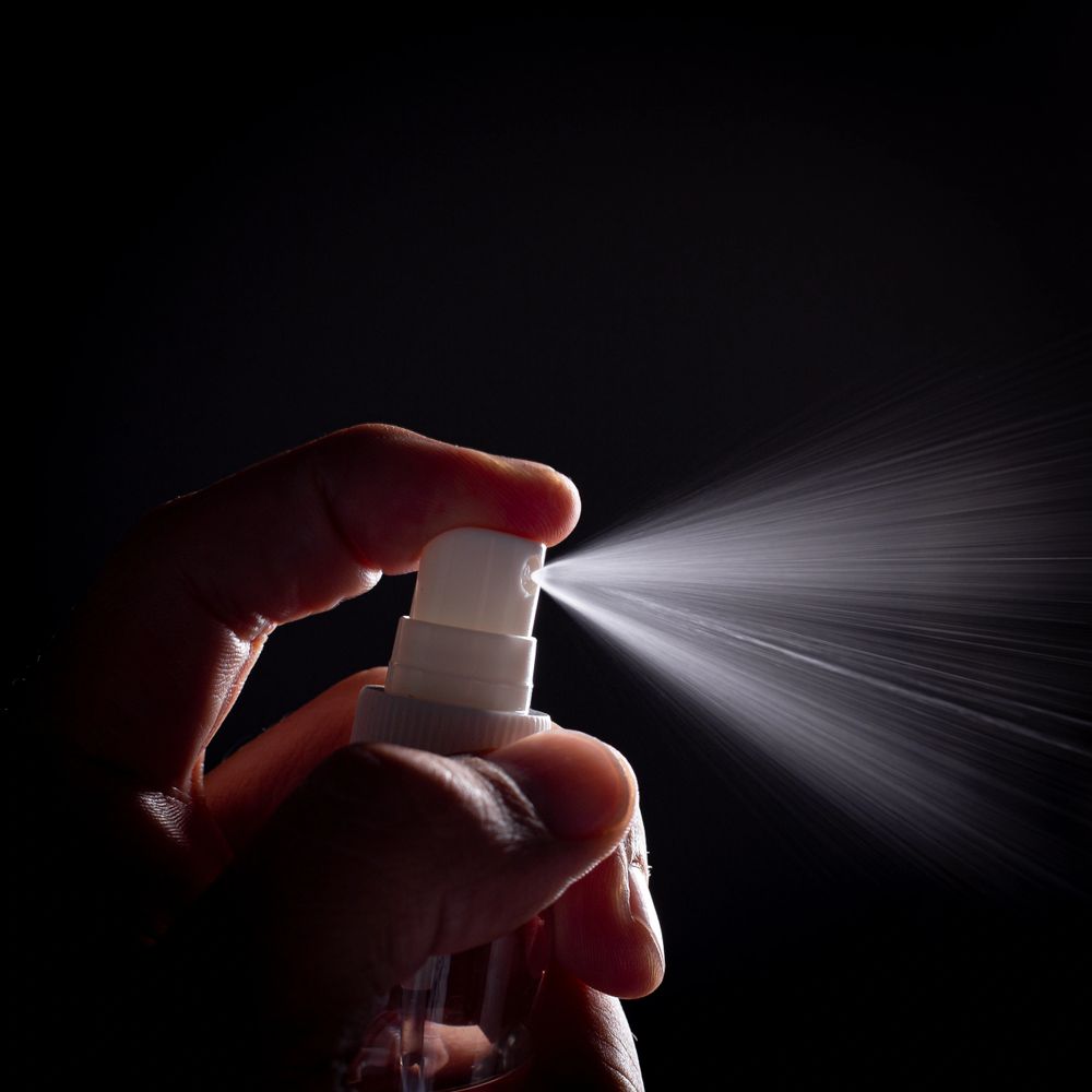 Hand spraying fine mist from a spray bottle against a dark background.