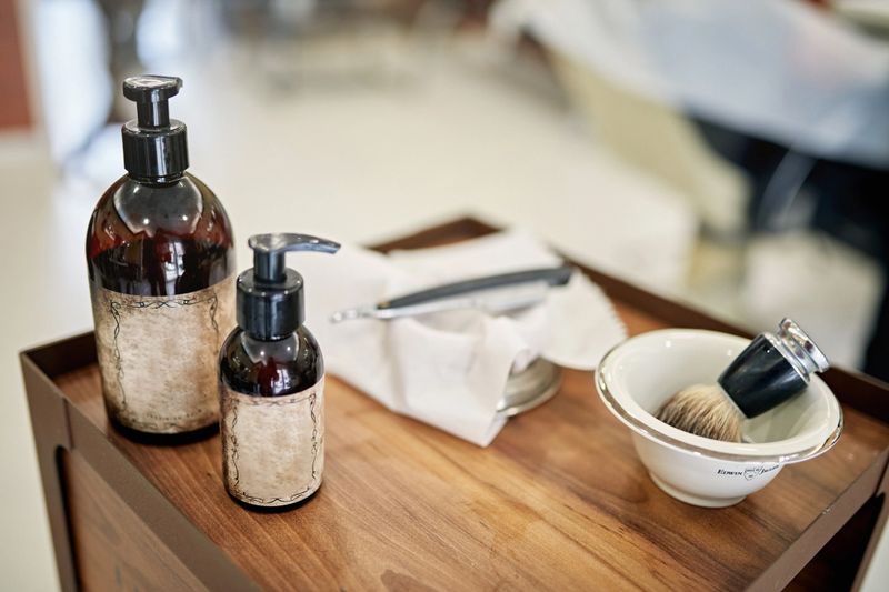 Close-up of shaving cream bowl with brush, oils, and other equipment on cart in professional barbershop and salon.