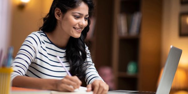 Young woman smiling while writing notes and looking at her laptop.